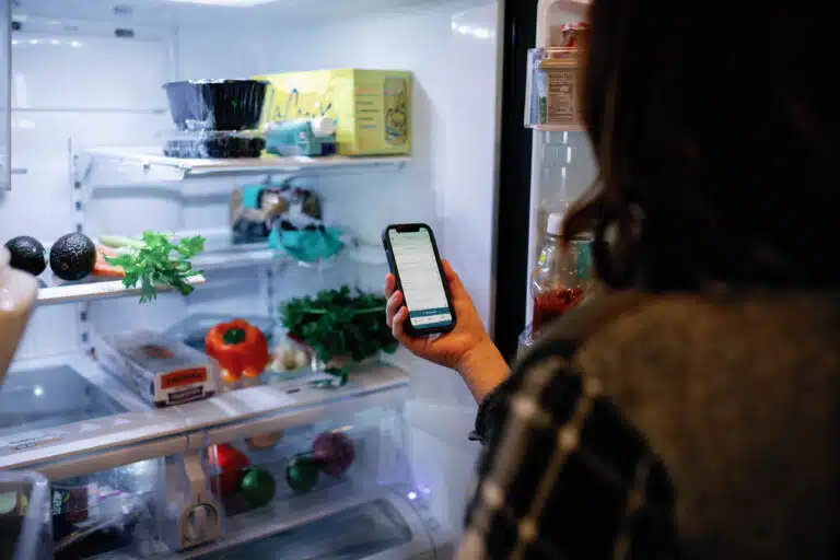 over the shoulder view of woman looking in her fridge with a phone shopping list in hand