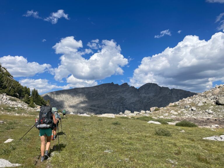 view of backpackers hiking across a mountain pass with a bright blue sky and peaks in the distance